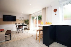 Un salon moderne doté d’un téléviseur à écran plat, d’un coin repas avec des chaises et une table, d’un comptoir de cuisine et d’une grande fenêtre agrémentée de rideaux.