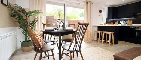 A dining area with a table and chairs, a kitchen area with a counter and stools, and a large window providing a view of greenery.