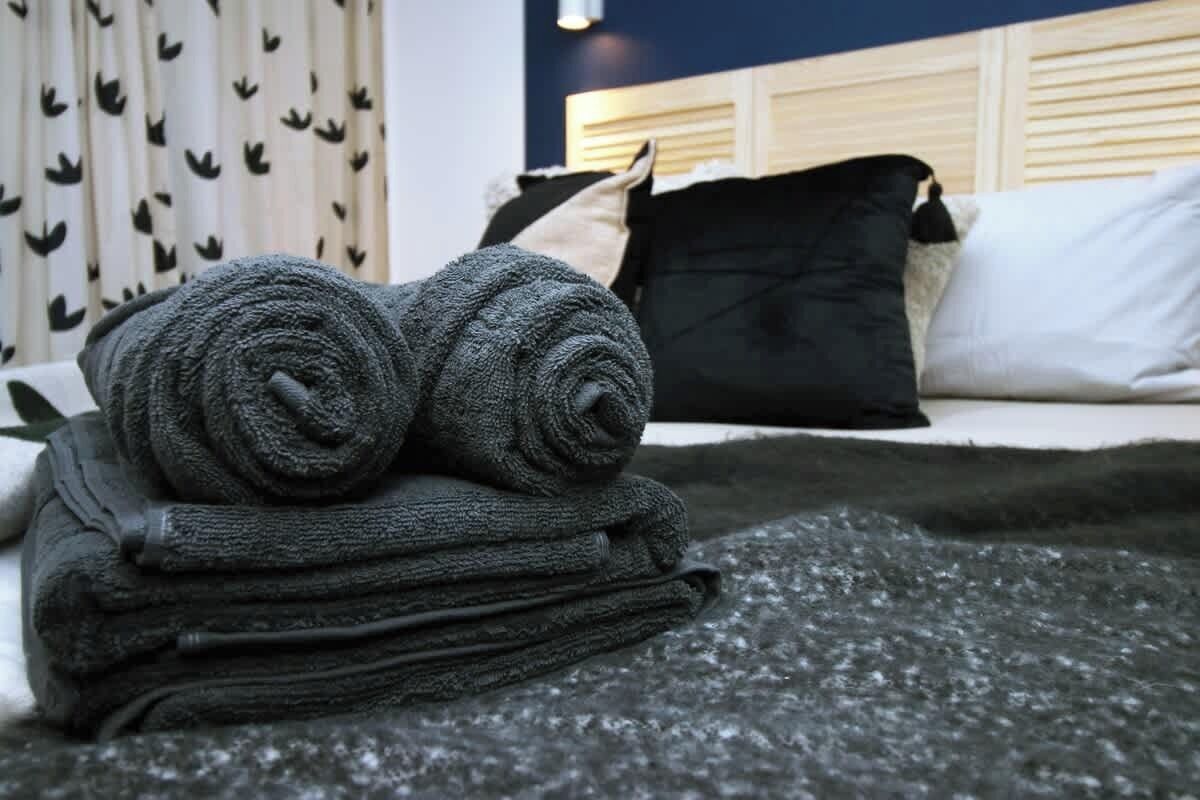 Un lit avec un oreiller blanc et noir, une couverture enroulée et un rideau orné d’un motif de feuilles.