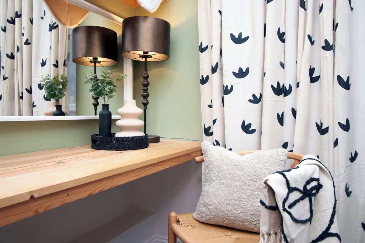 A room with a wooden bench, a white pillow, a blanket, two lamps, and a window with curtains.