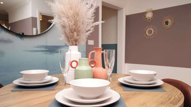 Una mesa de comedor redonda con platos y tazones blancos, un florero con plumas de pampa y espejos decorativos en la pared.