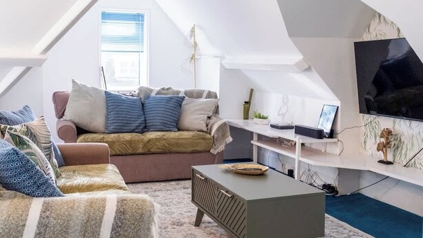 A modern loft-style room with a sloped ceiling, a sofa set, a coffee table, and a flat-screen TV mounted on the wall. - Boho Flat, Sleeps 8, Near Beach Ideal For Groups (Bournemouth)