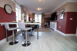 A modern kitchen with a dining area, a clock on the wall, and a TV mounted on the wall.