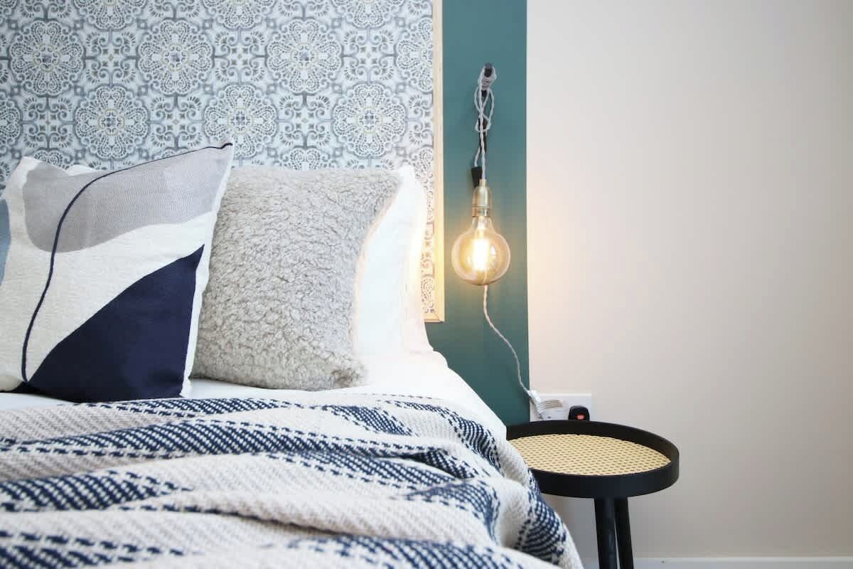 Un lit bien fait, avec une literie à motifs, un luminaire fixé au mur et une petite table ronde dont le plateau est fait d’un matériau tressé.