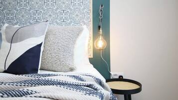 Un lit bien fait, avec une literie à motifs, un luminaire fixé au mur et une petite table ronde dont le plateau est fait d’un matériau tressé.