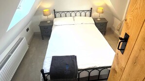A bedroom with a skylight, a round window, a bed, bedside lamps, a radiator, and a wooden door. - Small 3 bedroom converted stable in Norfolk (Upton)