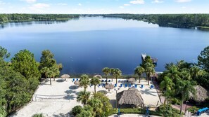 A beach with thatched umbrellas, lounge chairs, and a pier extending into a calm lake surrounded by trees.