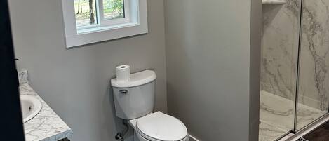 Et badeværelse med toilet, et vindue, en bruseniche med marmorvægge og trægulv.