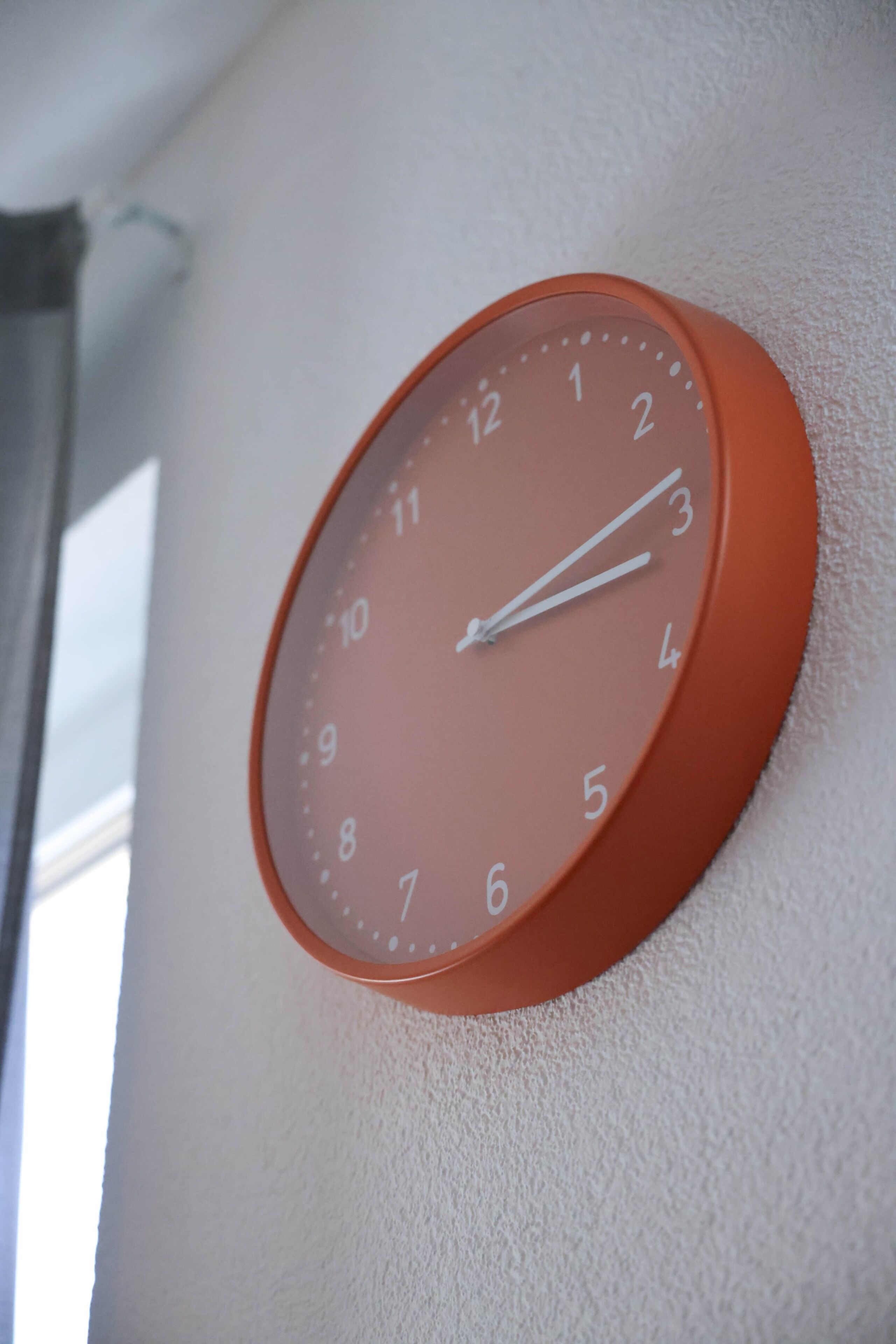 Eine runde, orangefarbene Wanduhr mit weißen Zahlen und Zeigern, an einer strukturierten Wand befestigt.