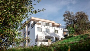A modern multi-story building with balconies, surrounded by greenery and trees.