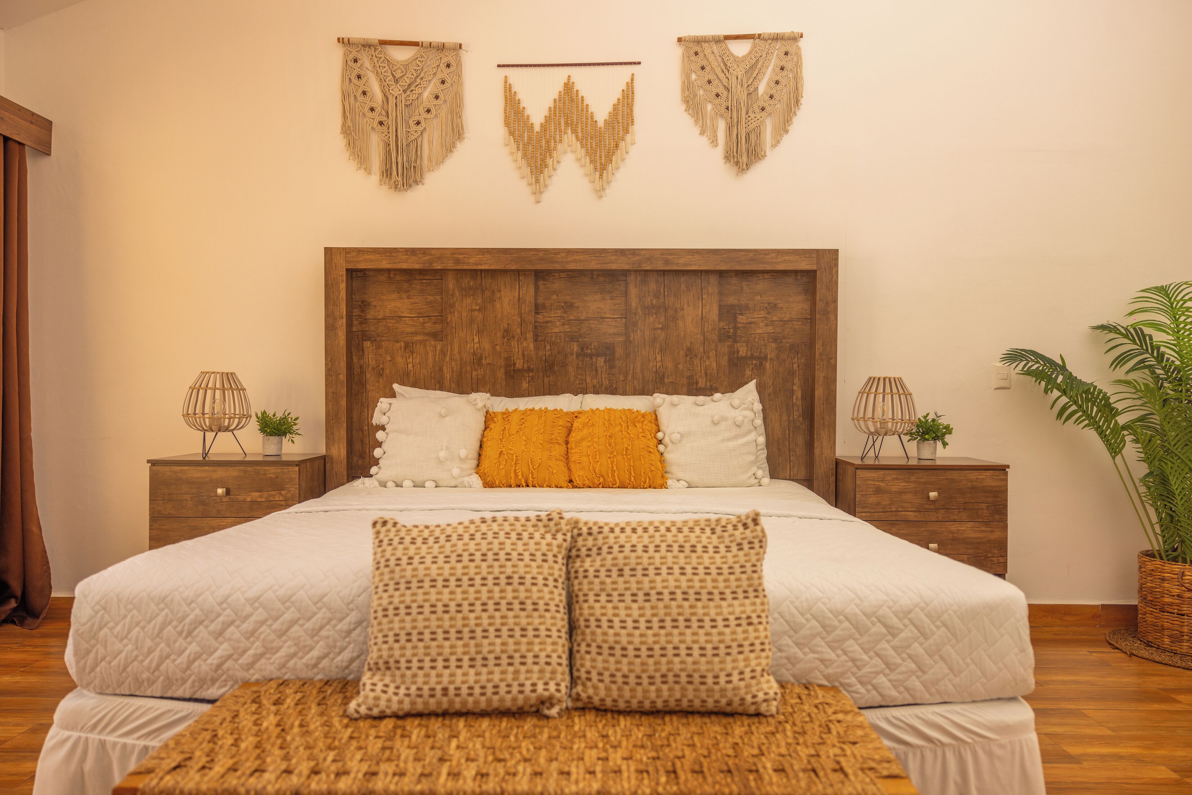 A bedroom with a wooden headboard, a white quilted bedspread, two patterned pillows, and a woven bench.