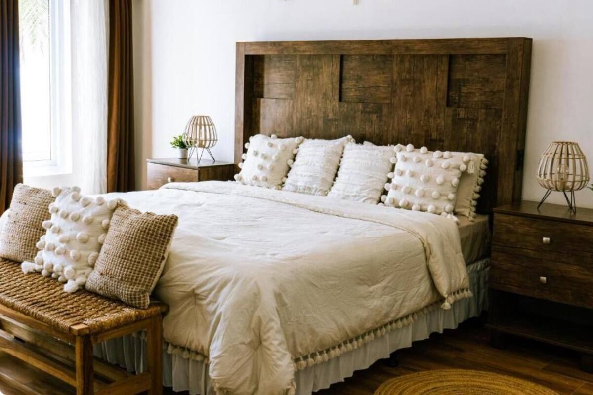 A bedroom with a wooden headboard, a large bed with white bedding and multiple pillows, a wicker bench, and a nightstand with a lamp.