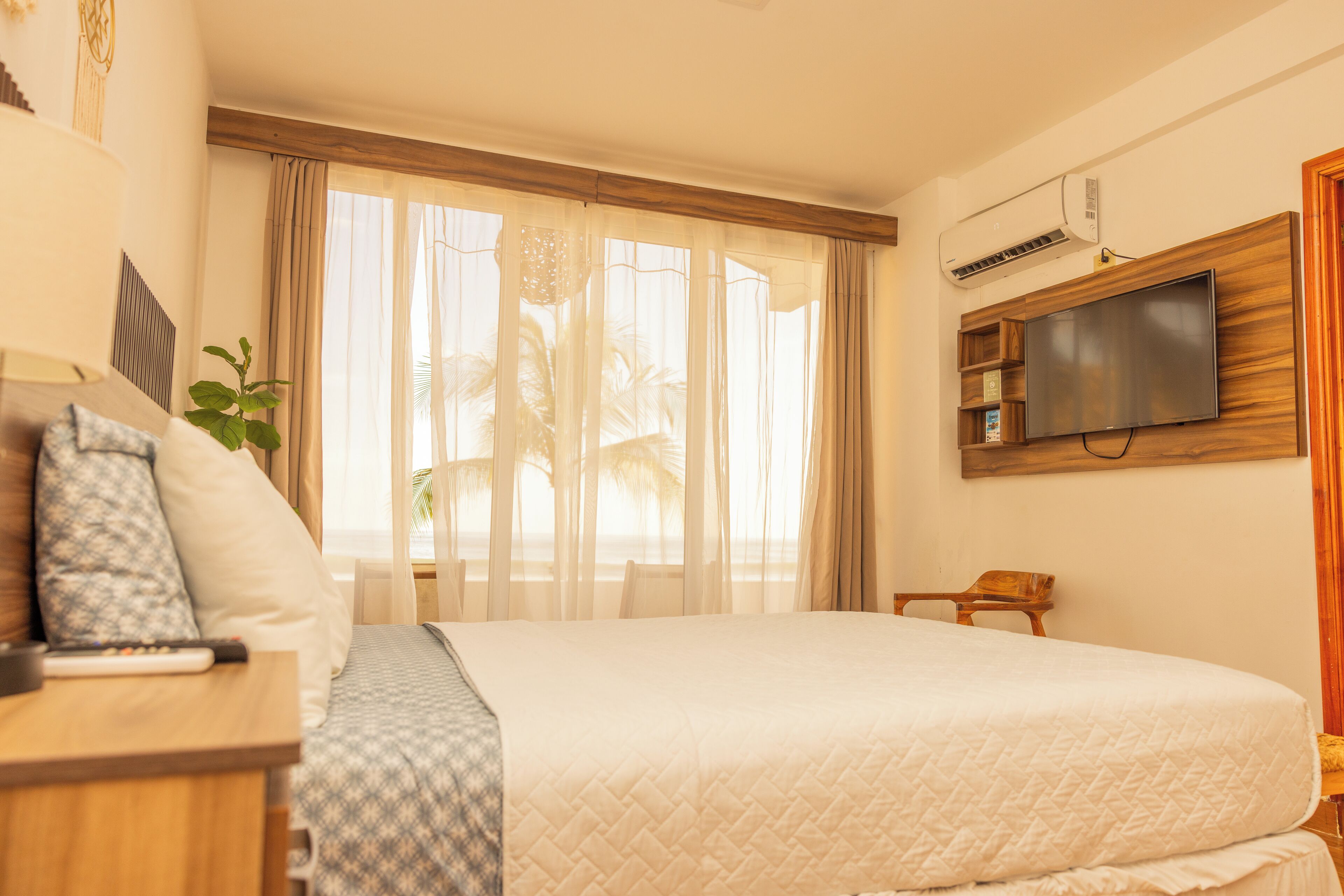 A hotel room with a large bed, a TV mounted on the wall, a bedside table with a remote control, and a window with sheer curtains.