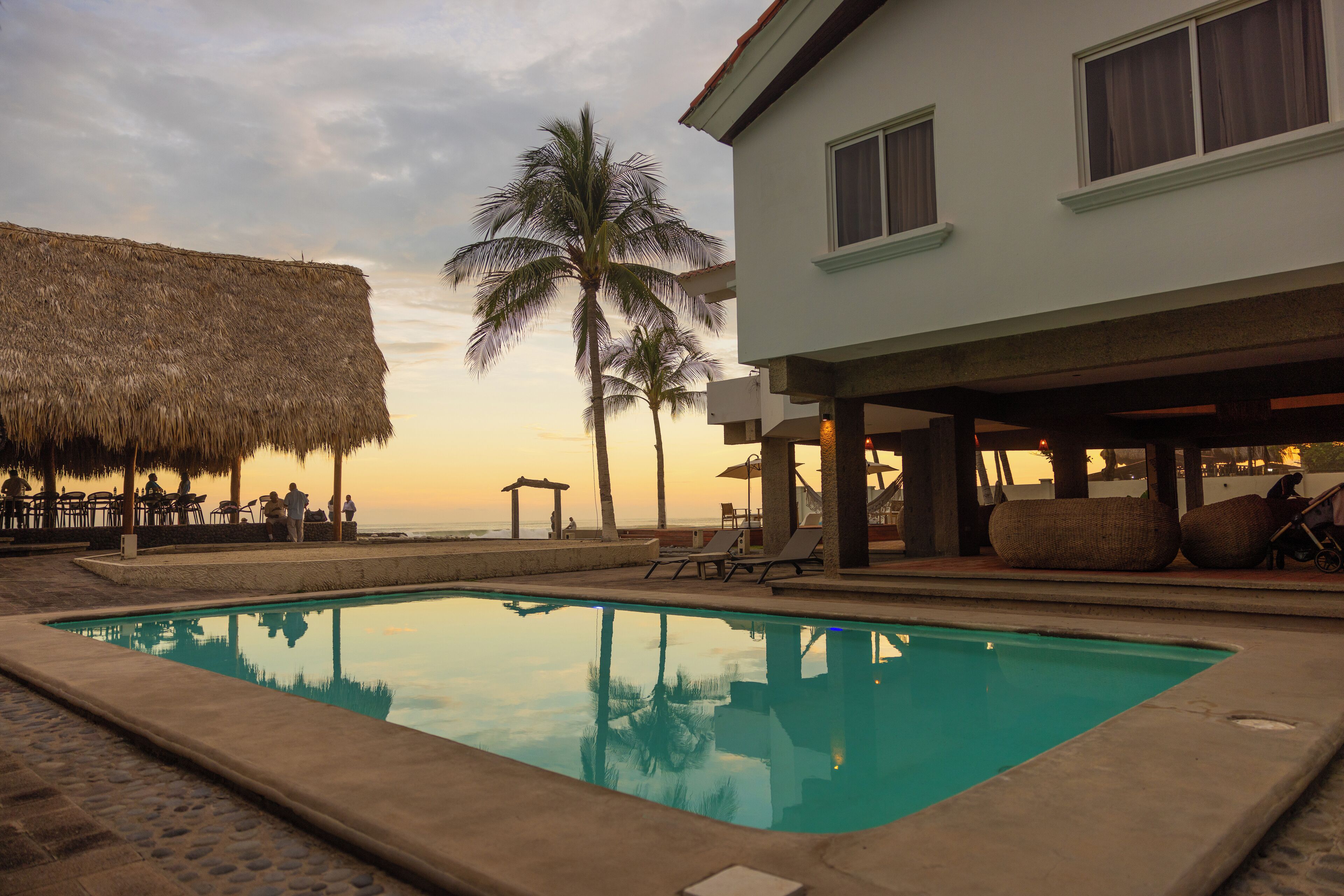A pool area with lounge chairs, a thatched roof structure, and a building with large windows.