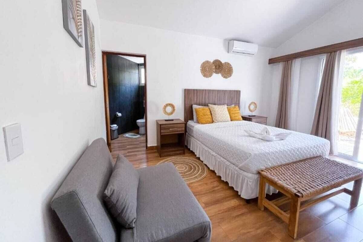 A bedroom with a bed, a bedside table, a bench, a sofa, and a door leading to another room.