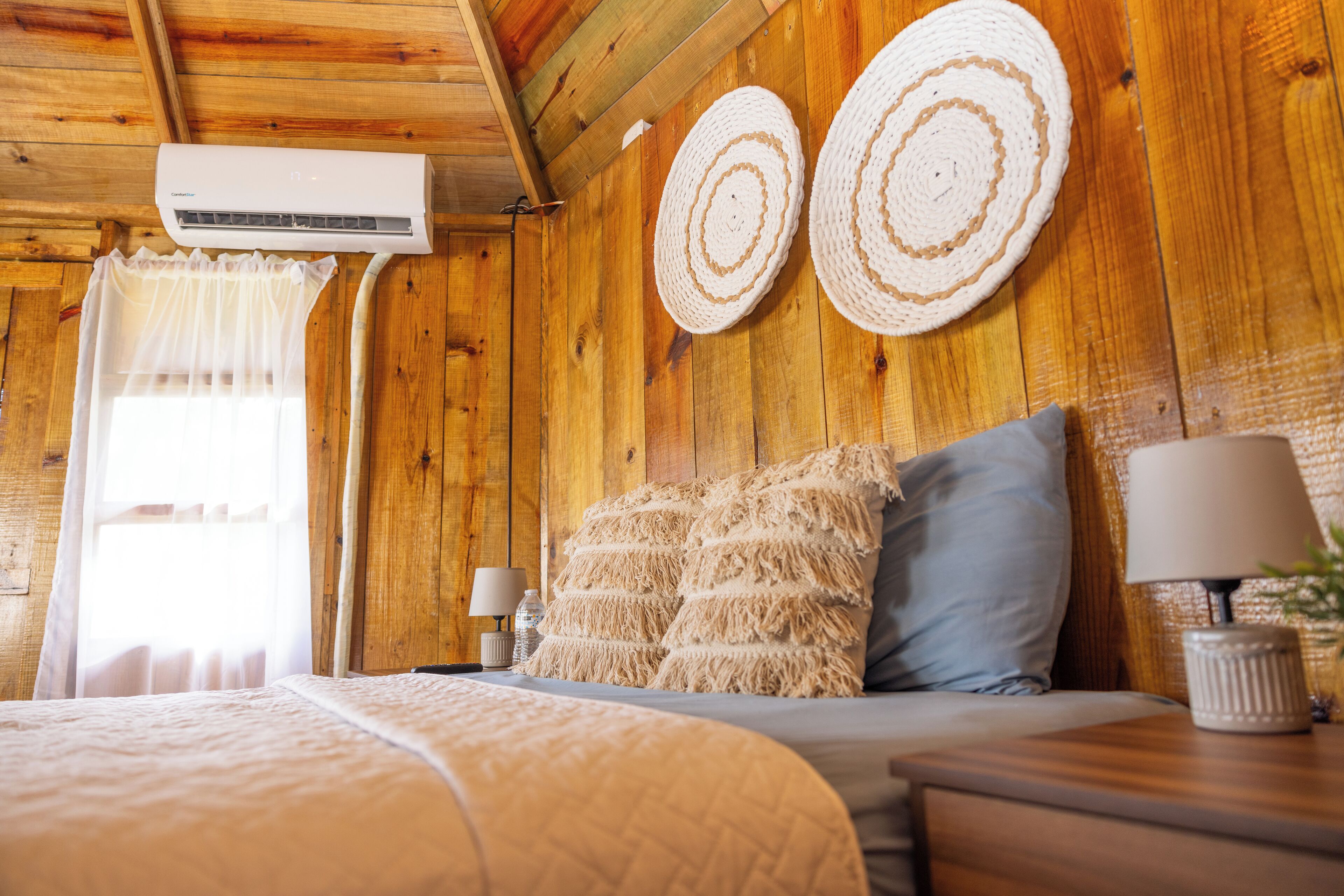 Habitación de madera con cama, dos tapices tejidos, una lámpara de noche y un equipo de aire acondicionado.