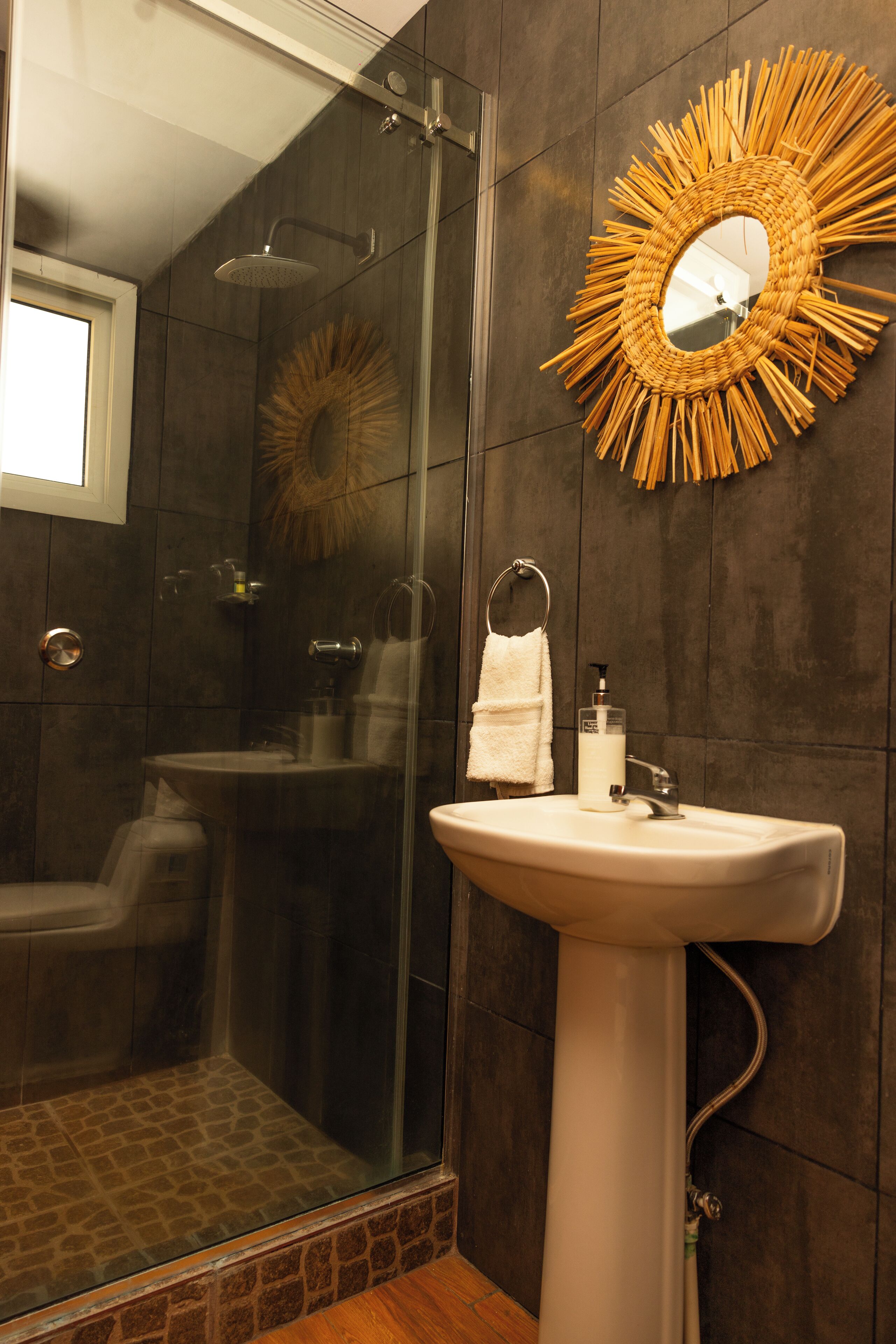 A bathroom with a glass-enclosed shower, a white pedestal sink, a mirror with a sunburst frame, and dark wooden walls.