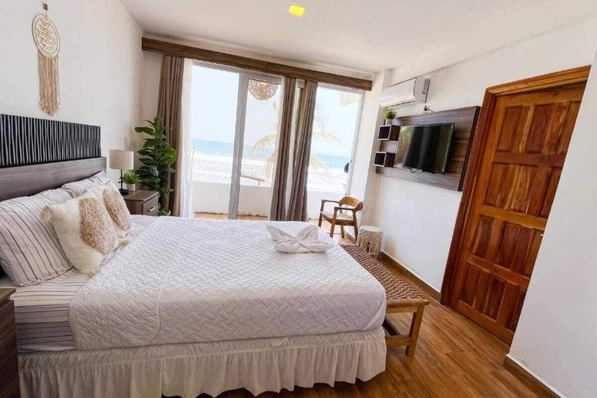 A bedroom with a bed, a TV, a wooden door, and a view of the beach.