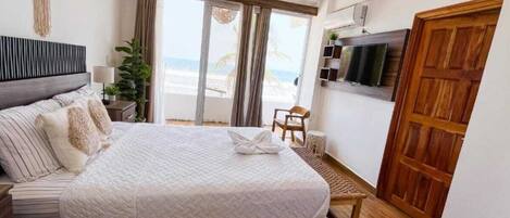 A bedroom with a bed, a TV, a wooden door, and a view of the beach.