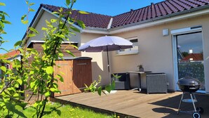 Un patio trasero con una terraza de madera, una parrilla, una mesa con sillas y un portón corredizo de vidrio que da a un patio.