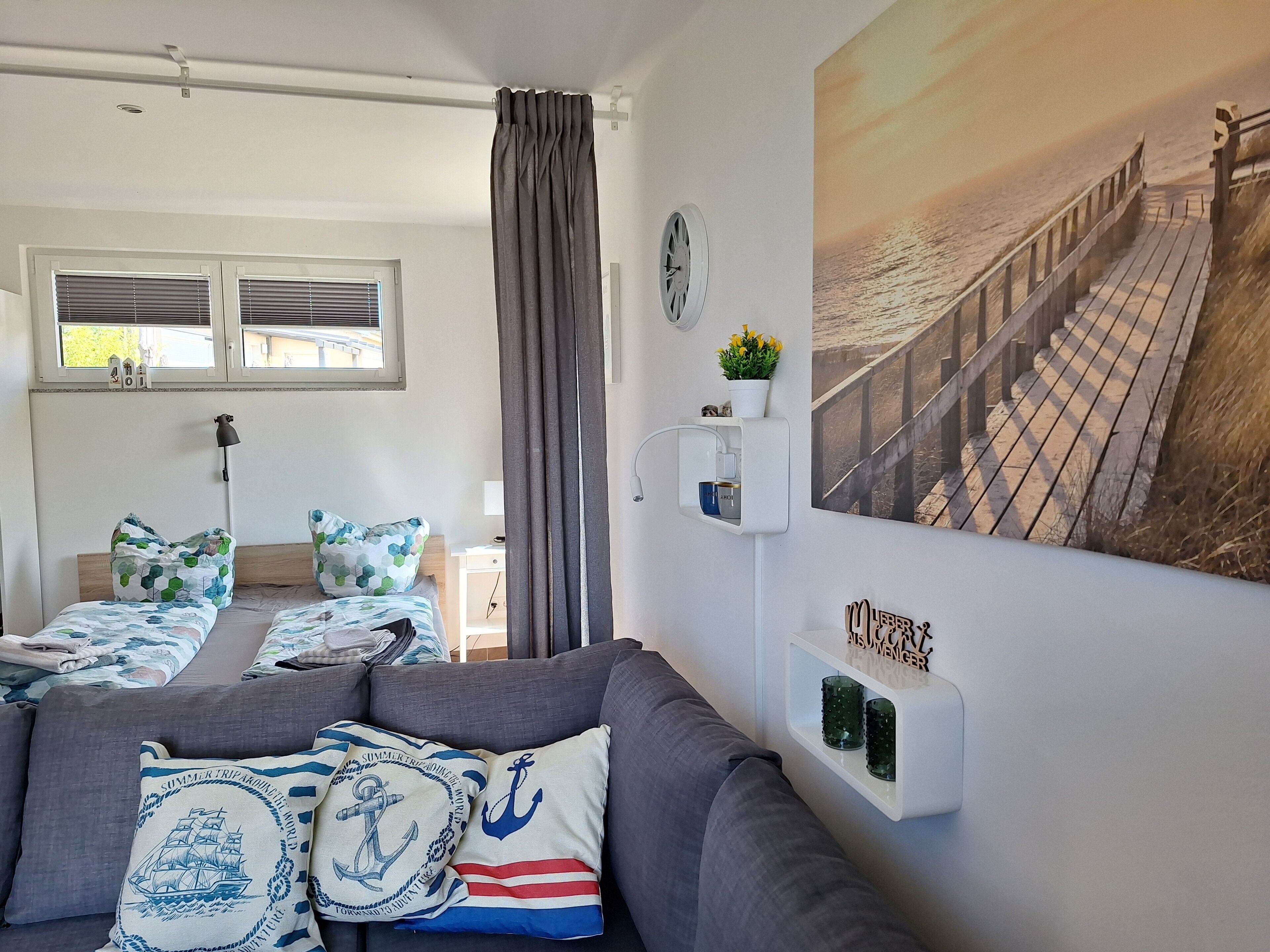 Una habitación con un sofá gris, una cama con ropa de cama estampada, un estante montado en la pared con objetos decorativos, una ventana con persianas y una fotografía grande de una playa.