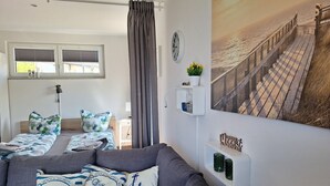 Una habitación con un sofá gris, una cama con ropa de cama estampada, un estante montado en la pared con objetos decorativos, una ventana con persianas y una fotografía grande de una playa.