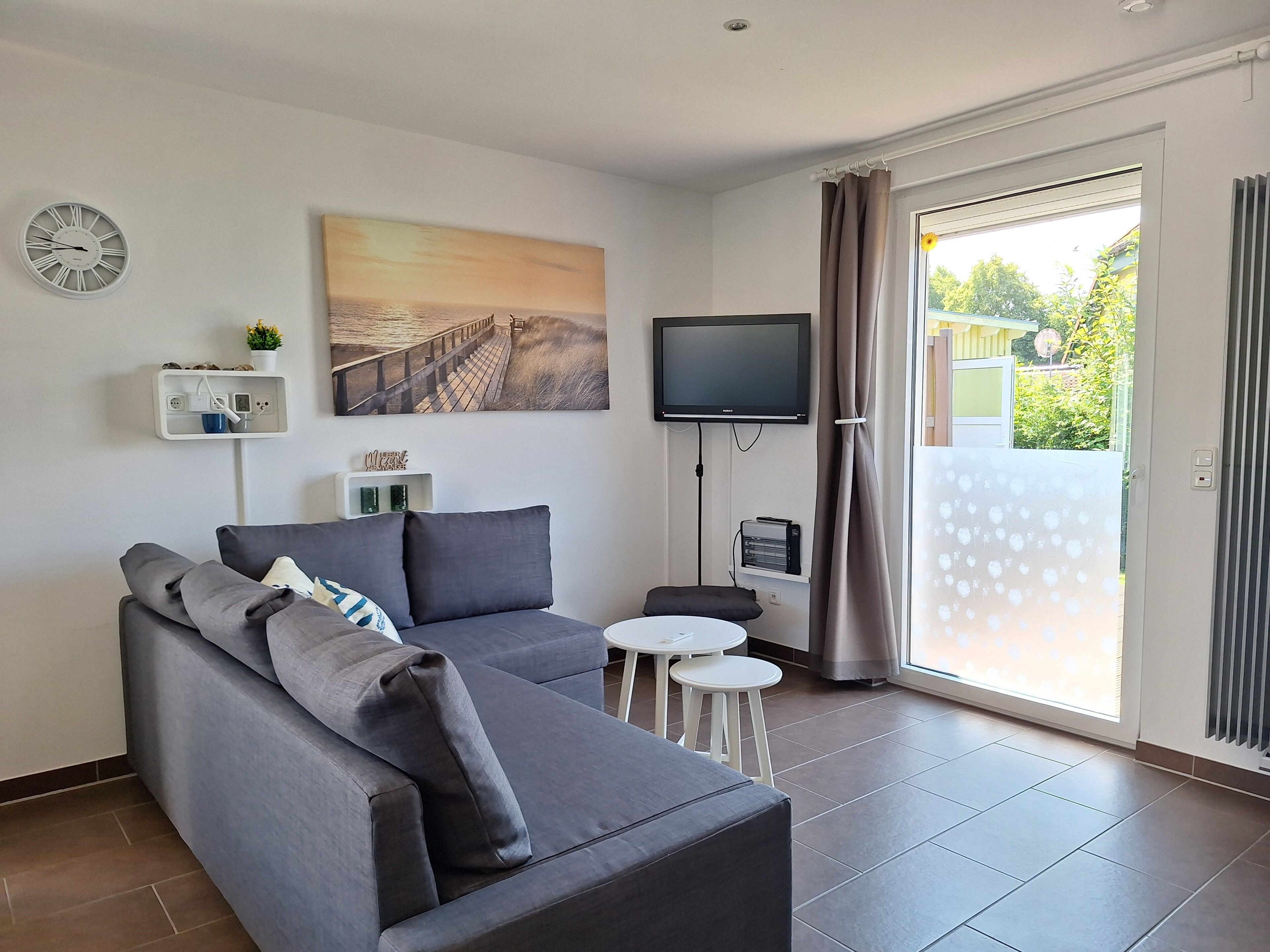 Un salon moderne avec un canapé gris, un téléviseur à écran plat fixé au mur, une petite table ronde et une porte-fenêtre coulissante donnant sur un espace extérieur.