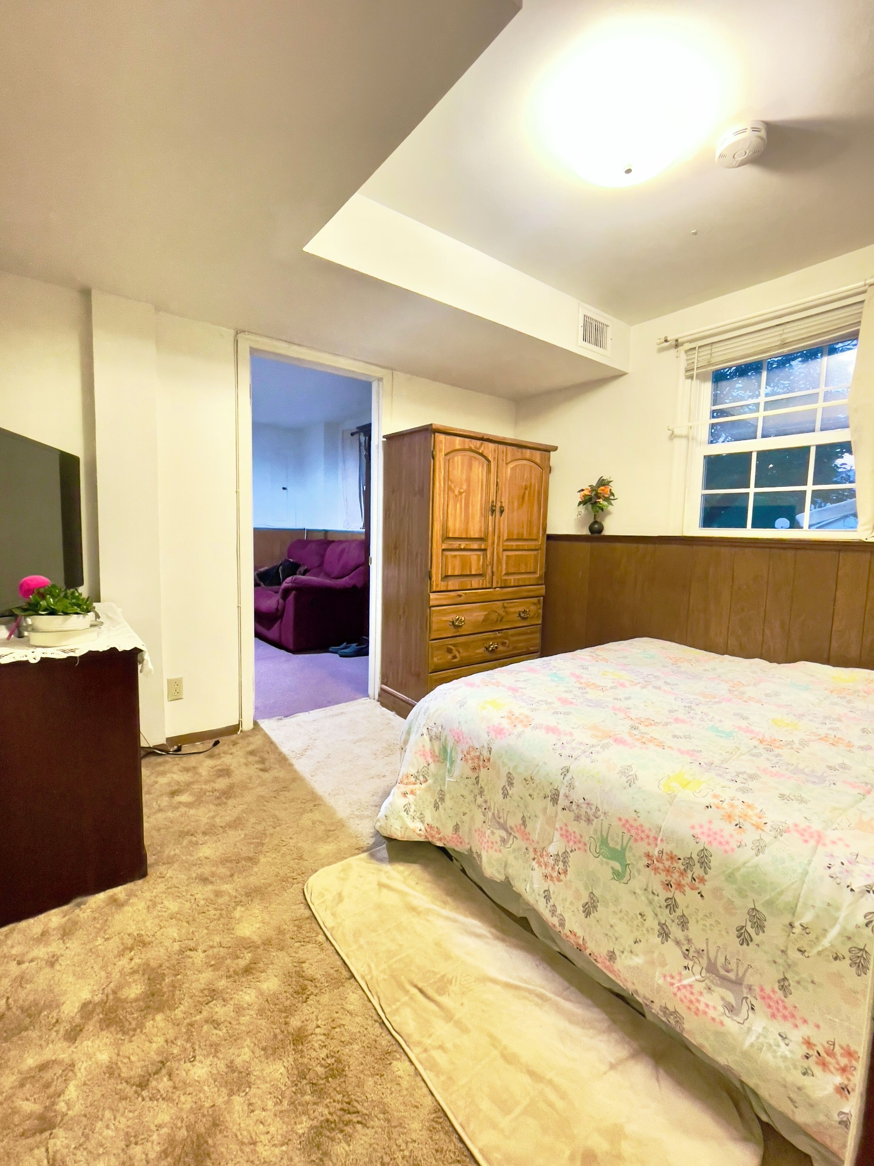 A bedroom with a bed, a dresser, a wardrobe, a window with blinds, and a door leading to another room.