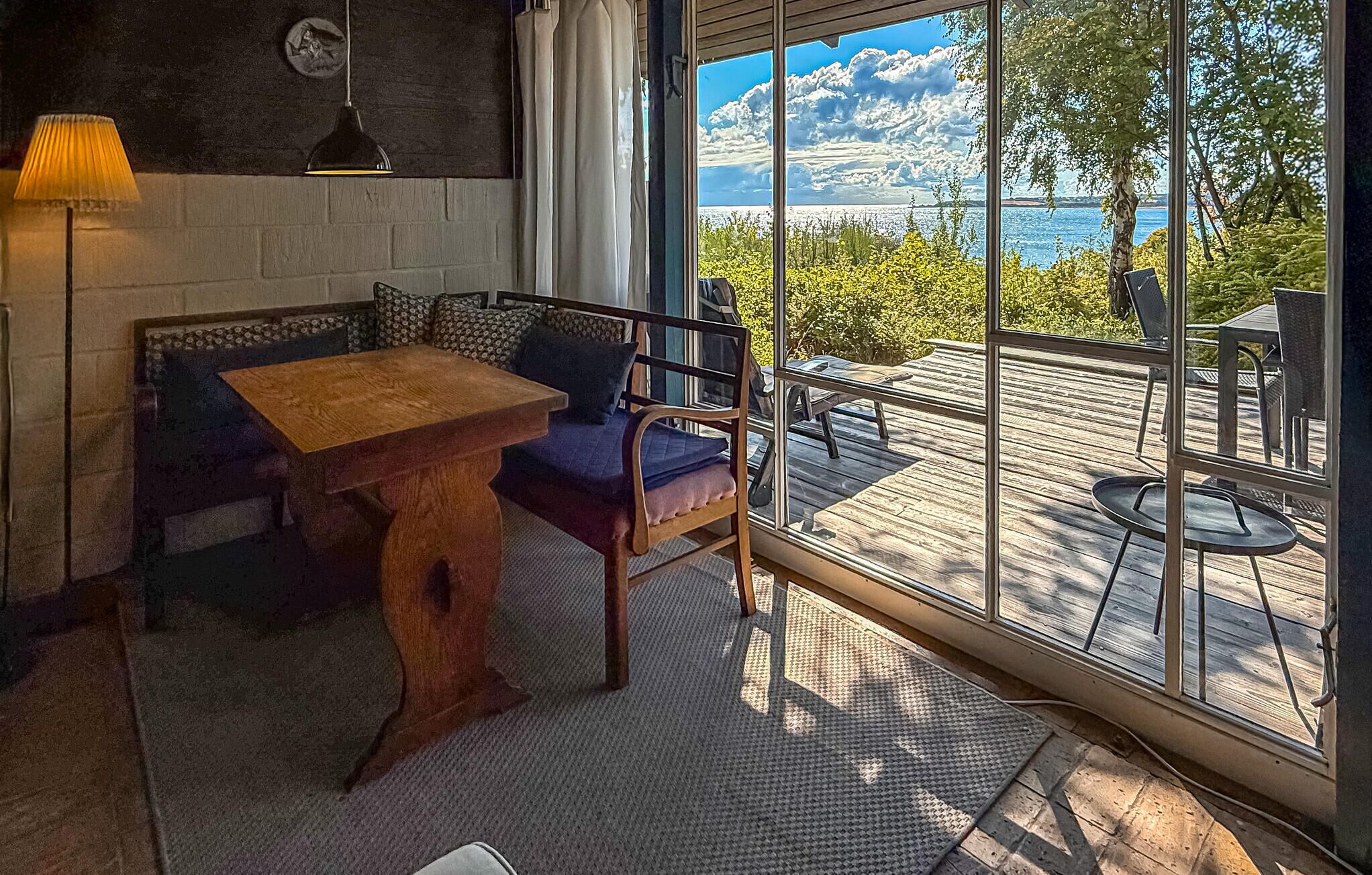 A cozy room with a wooden table and blue sofa, a large window with a view of the sea, and a balcony with outdoor furniture.