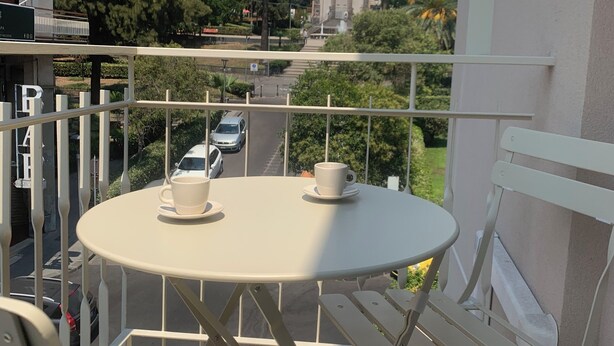 A balcony with a round table and two chairs, overlooking a street and a building with a statue. - Etna Guest House (Belpasso)