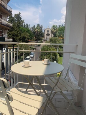 A balcony with a round table and two chairs, overlooking a street and a building with a statue. - Etna Guest House (Belpasso)