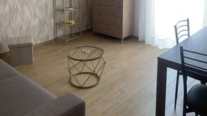 A modern living room with a flat-screen TV mounted on the wall, a wooden herringbone-patterned wall, a glass coffee table, a dining table with chairs, and a small stool. - Etna Guest House (Belpasso)