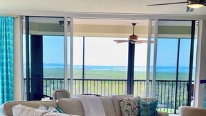 A spacious living room with a large white sofa, a round ottoman, and a view of the ocean through large windows.