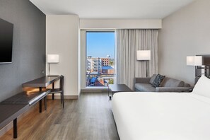 A hotel room with a bed, a desk, a chair, a sofa, a TV, and a window with a city view. - AC Hotel Auburn (Auburn)