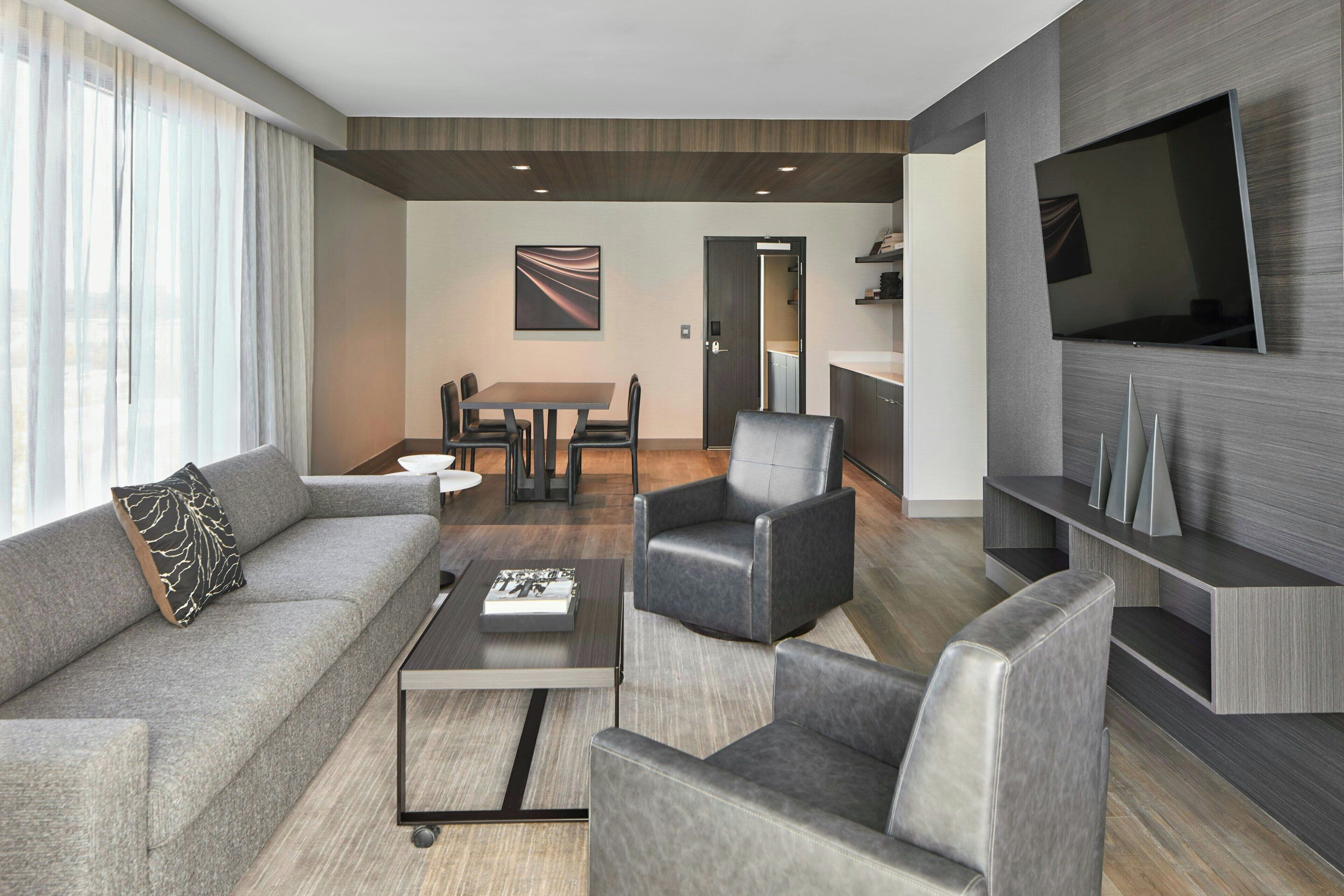 A modern living room with a sofa, armchairs, a coffee table, a TV, and a dining area in the background.