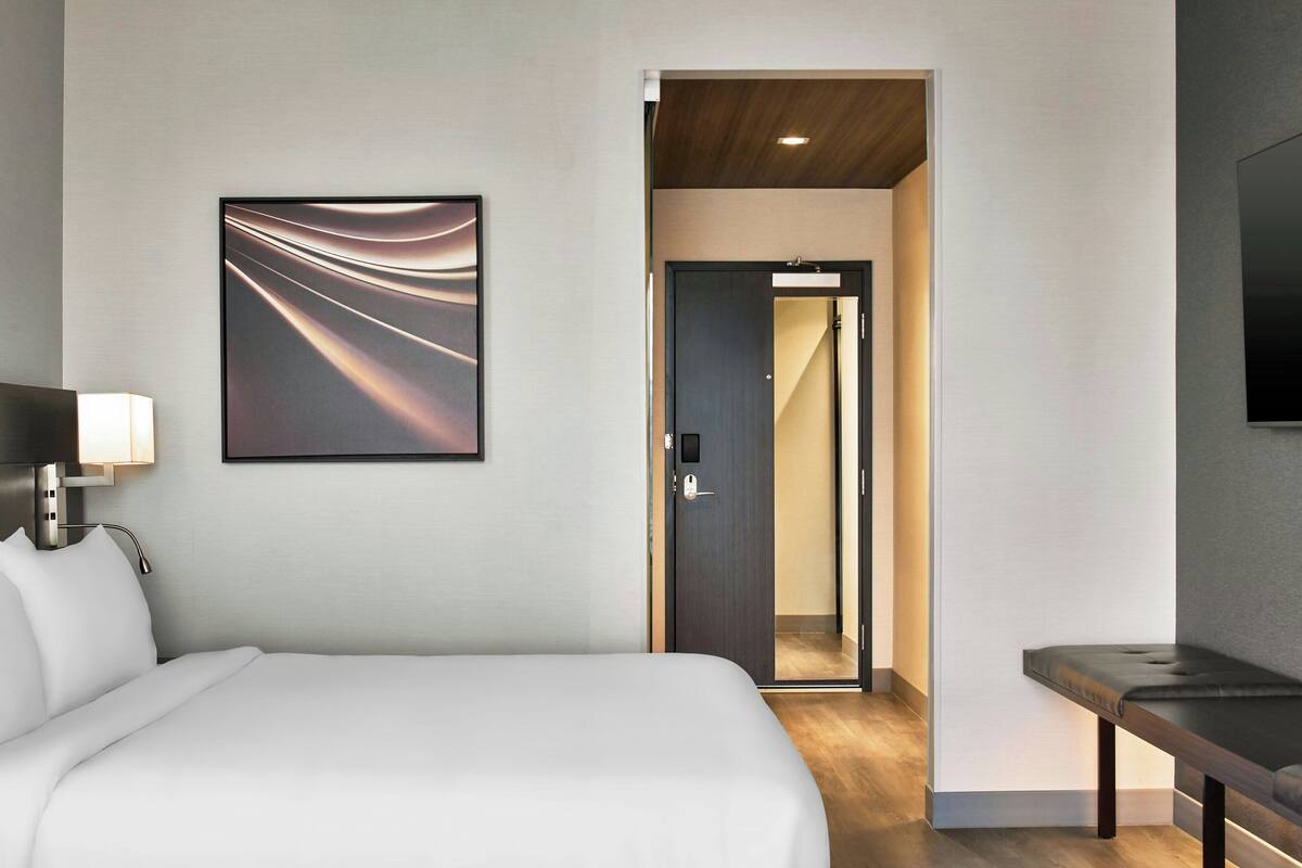 Quarto de hotel com cama, mesa de cabeceira, televisão na parede e porta para o banheiro.