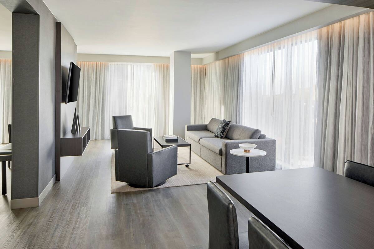 Quarto de hotel moderno com sofá, cadeiras, mesa de jantar e televisão fixada na parede.