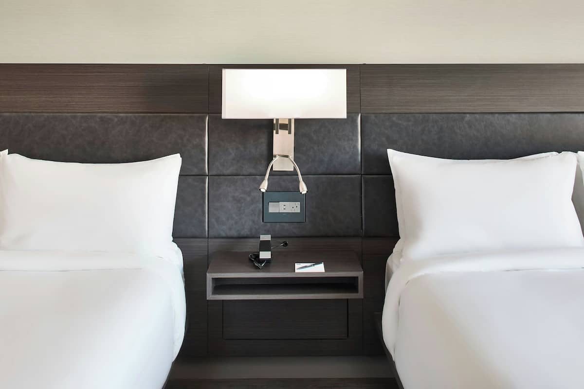 Quarto de hotel com duas camas, uma mesa de cabeceira com abajur e uma tomada.