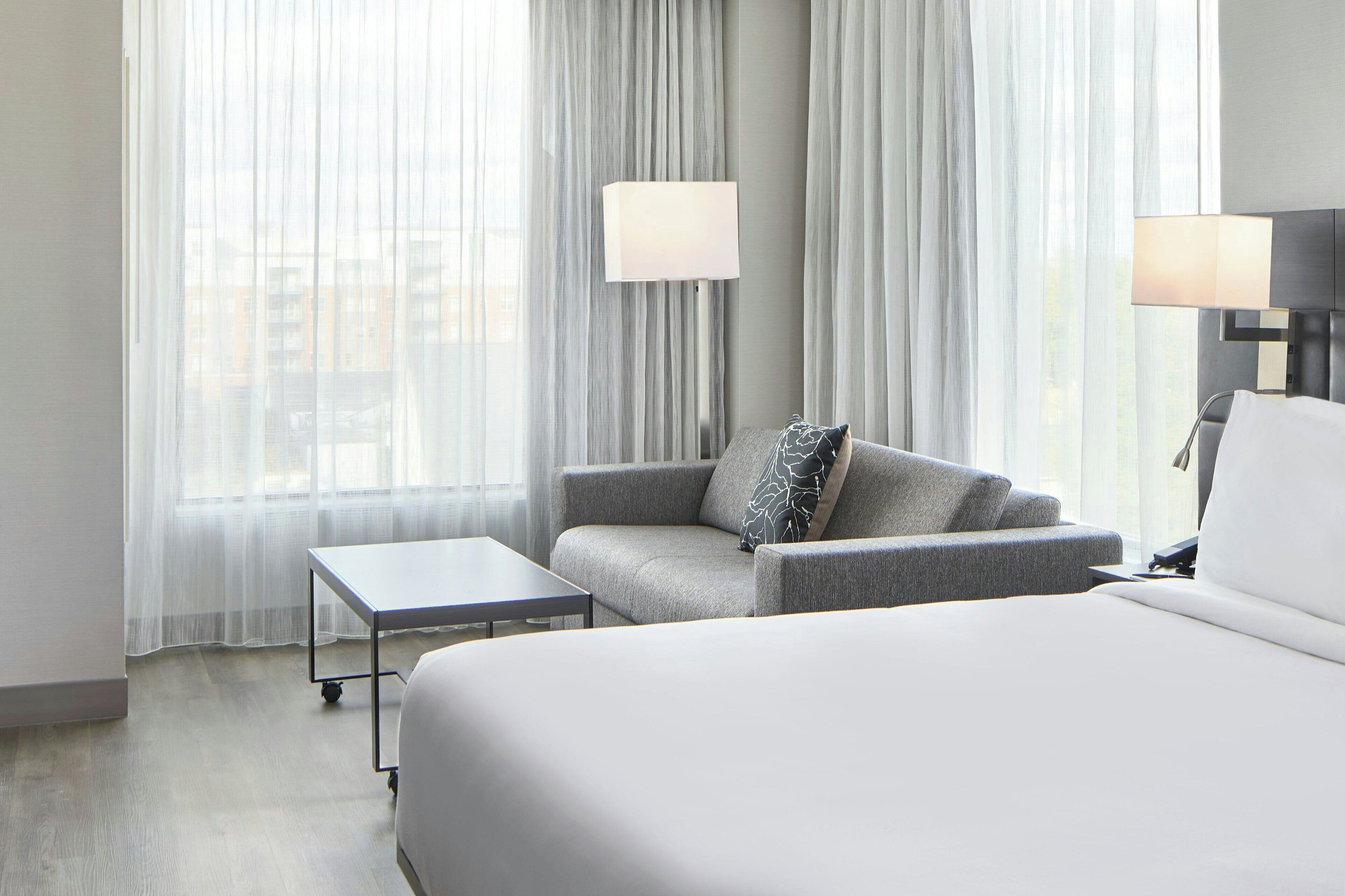 A hotel room with a large bed, a grey sofa, a small table, two wall-mounted lamps, and sheer curtains.