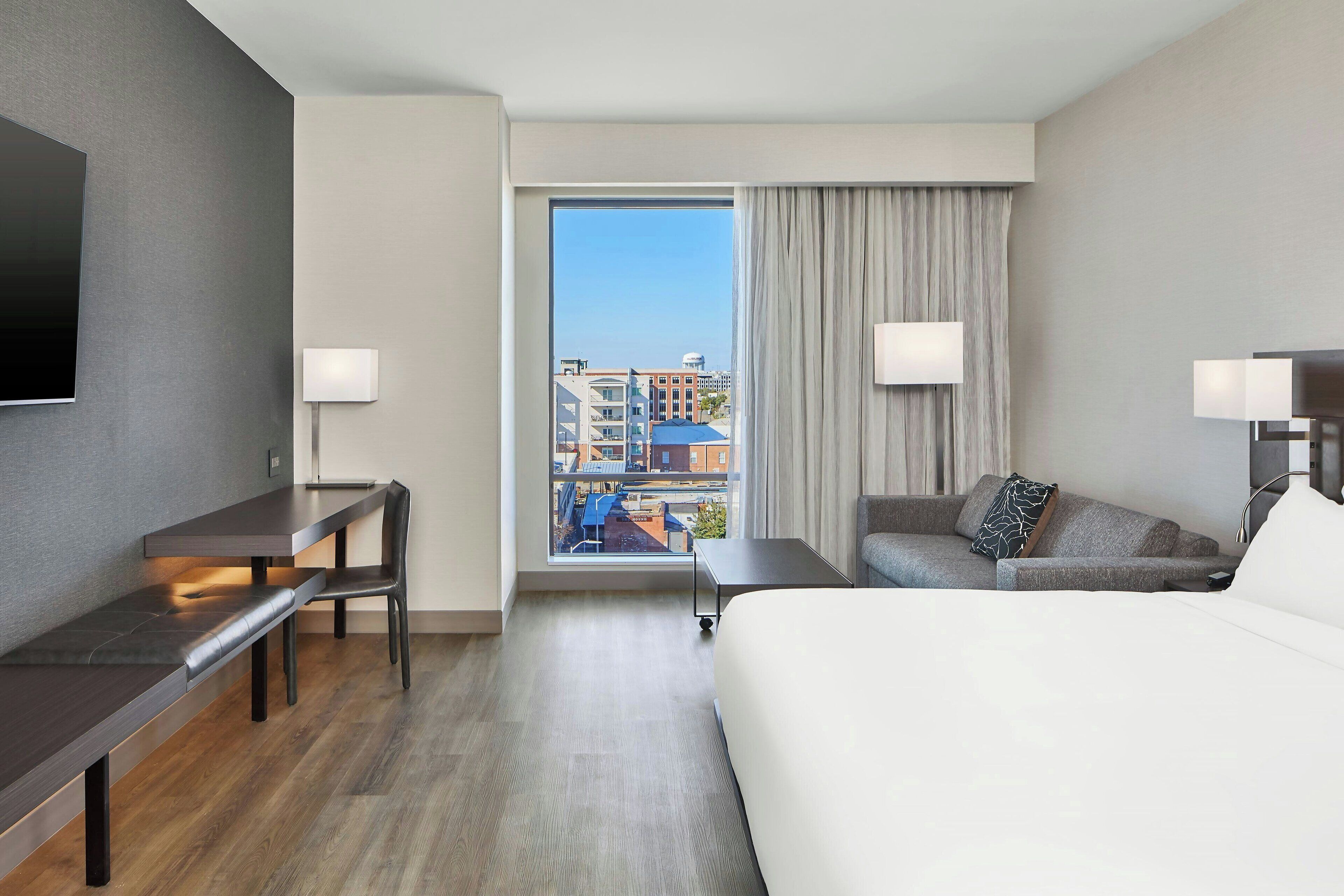 A modern hotel room with a large bed, a desk, a chair, and a sofa. There is a large window with a view of buildings and a water tower.