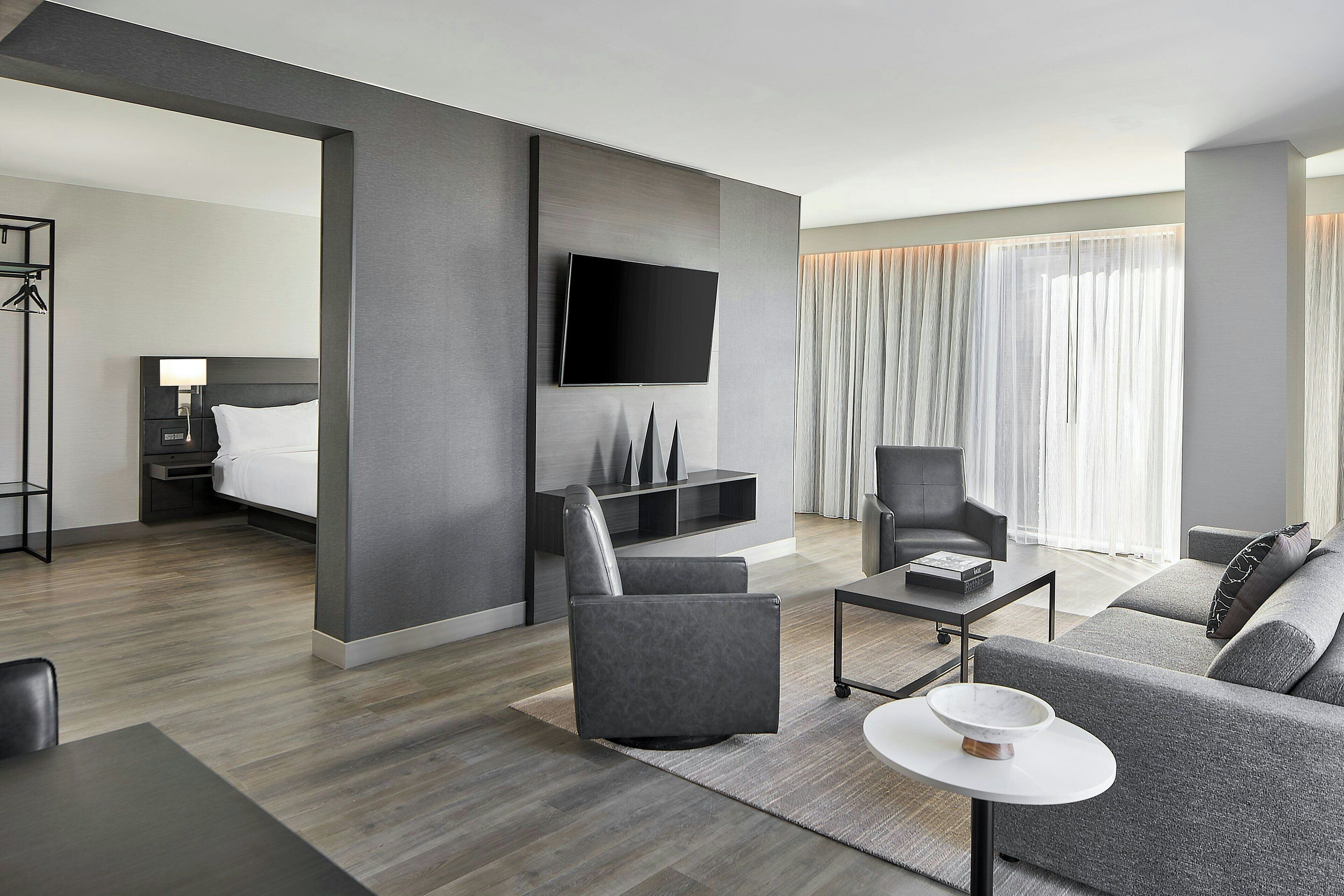A modern hotel room with a grey sofa, a flat-screen TV mounted on the wall, a small table with a bowl, and a bed with a white headboard.