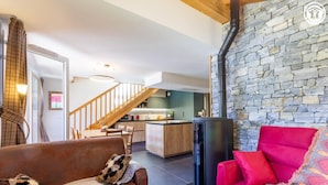 A cozy living room with a brown leather sofa and a red armchair, a stone wall, and a kitchen in the background.