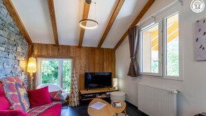 A cozy room with a stone wall, wooden beams, a TV, a fireplace, and a window with curtains.