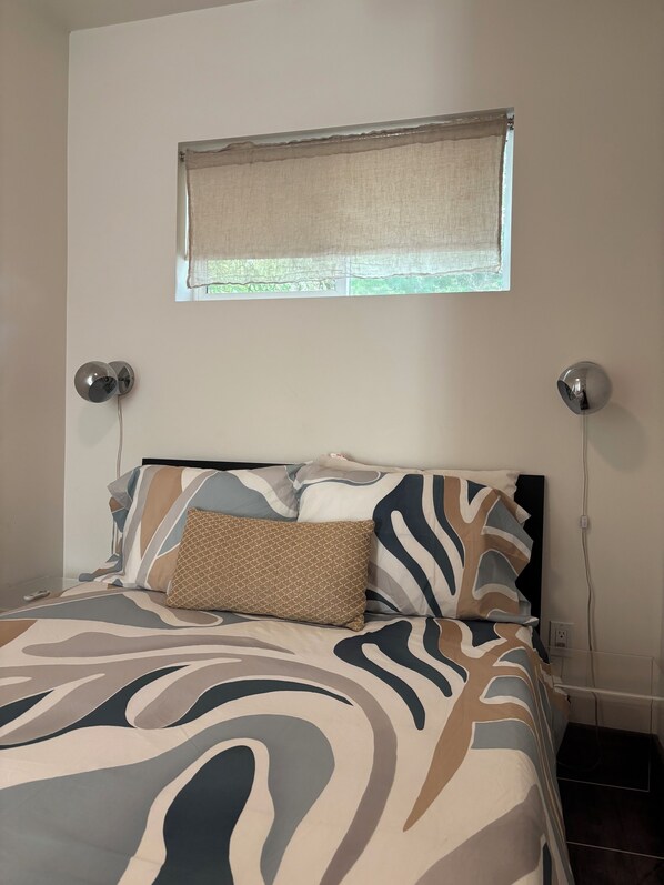 A bedroom with a bed, a window with a curtain, two wall-mounted lamps, and a power outlet.