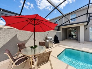 Un espace aménagé au bord de la piscine, avec une table et des chaises en verre, un parasol rouge et un barbecue.