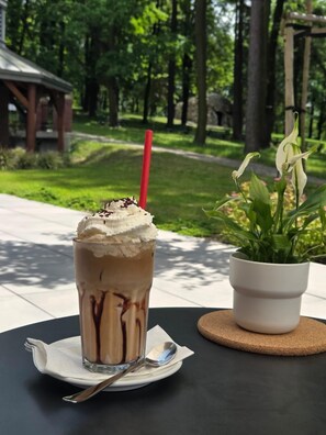 Un vaso alto de café helado con crema batida y jarabe de chocolate, una pajita roja y una cuchara sobre un paño blanco. Un pequeño macero blanco con plantas verdes está sobre un posavasos de corcho junto al vaso.