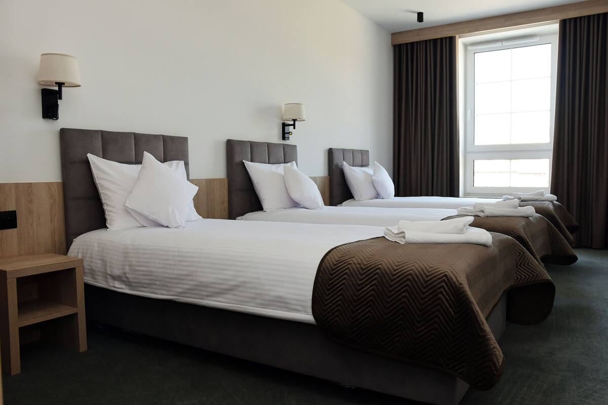 A hotel room with three beds, each with white bedding and brown headboards. There are wall-mounted lamps on either side of the beds.