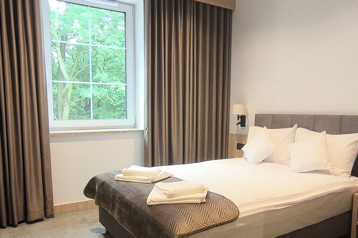 A modern hotel room with a large bed, a window with curtains, a headboard, and a bedside table with a lamp.