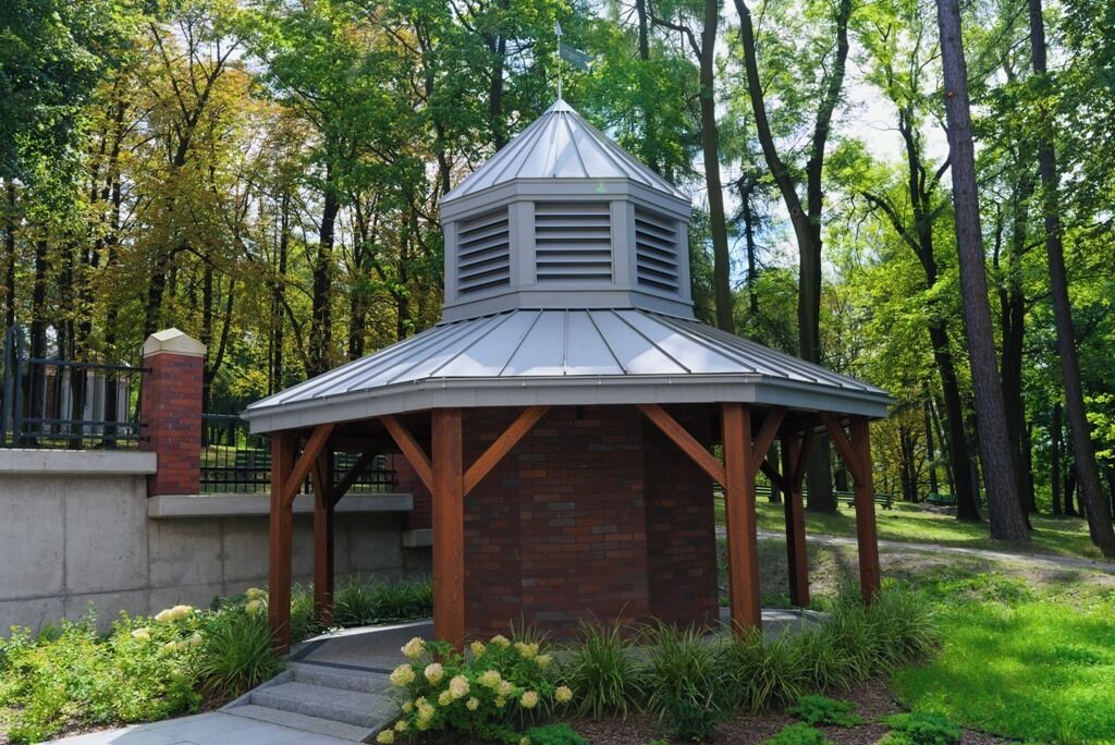 A gazebo with a metal roof and brick base, surrounded by greenery and trees.