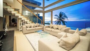 A modern living room with a large glass wall overlooking a beach at dusk.